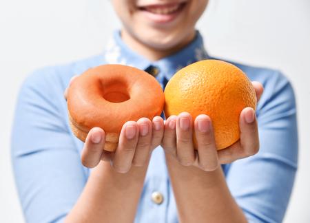 Dieting concept. Beautiful Young Asian Woman in blue shirt holding orange and chocolate donut on white background.
Choosing between junk food with healthy food.の写真素材
