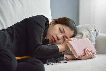 Exhausted, Tired Asian Business woman in black shirt holding notebook and sleeping on white sofa. Stress from overtime working concept.の写真素材