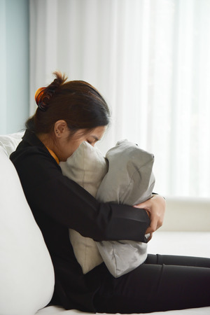 Exhausted, Tired Asian Business woman in Yellow shirt and black suit sitting on white sofa and hiding her face on pillow with blue wall. Stress from overtime working concept.の写真素材