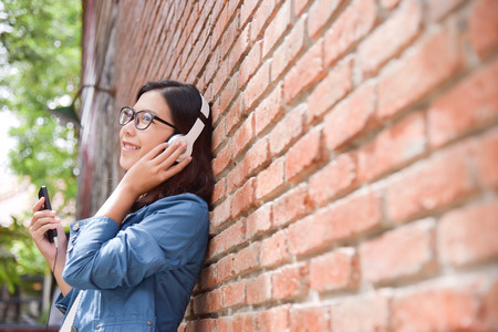 Happy Beautiful Asian woman in blue shirt listening to the music with headphone via her smart phone on old red brick wall background. copy space.の写真素材
