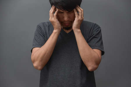 Stressed Asian beard man in Gray shirt standing and having headache on grey background. Migraine from Overtime working.の写真素材
