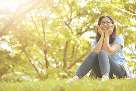 Happy Asian woman with Glasses in Blue shirt sitting on grass and smiling with Green natural background. Girl with Outdoor park. Copy space.の写真素材