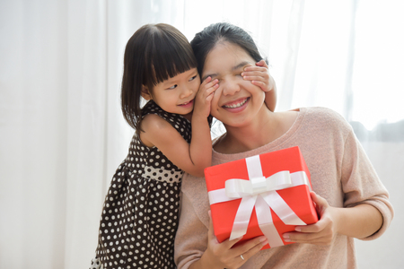 Making a surprise for Birthday, Christmas and New year. Asian Cute little girl covering her mother eyes with her hands while giving a red gift box to her on white background.の写真素材
