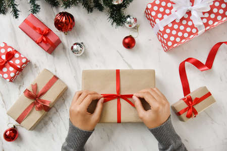 Female hands wrapping a brown present box with red ribbon on white marble background for Christmas and New year. Top view.の写真素材