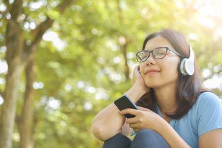 Happy Asian Young beautiful woman in Glasses listening music with headphones and smart phone at Park outdoor. Smiling face. Copy space.の写真素材