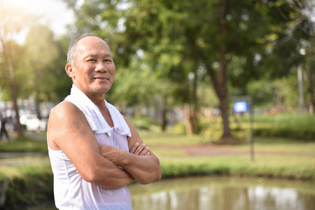 Confident Asian Senior male posing with arms crossed and smiling while exercising at park outdoor background.の写真素材