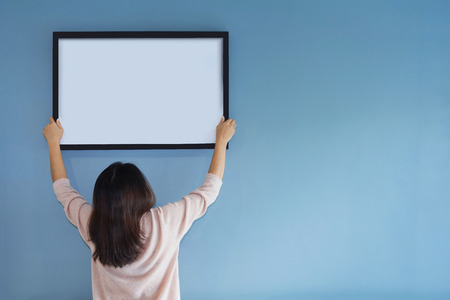 Asian woman holding a picture frame on blue wall. Rear view. Copy space.の写真素材