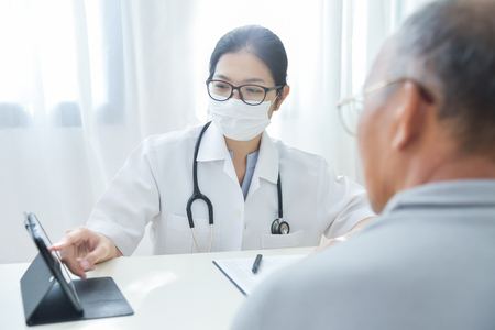 Asian Female Doctor with protective mask discussing with senior man patient about a chest radiograph in digital tablet computer in medical office. X-ray.の写真素材