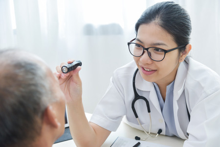 Young Asian Female Doctor wear glasses checking senior man patient eyes with flashlight in medical office.の写真素材