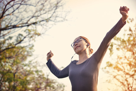 Happy Asian female in grey sportswear workout at the park. Young woman is warming up and stretching her arms at outdoors on morning. Health care and sport concept. Listening to the music with Earphones. Copy space. Exercising.の写真素材