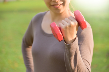 Asian female in grey sportswear is lifting dumbbell at park. Young woman is warming up outdoors on morning. Health care and sport concept. Listening to the music with Earphones. Sunset and Sunlight. Copy space. Exercising.の写真素材