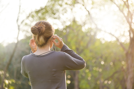 Happy Young Asian woman in Grey sportswear is smiling and listening to the music with earphones at outdoors on morning. Female workout at the park. Health care concept.  Sunset and Sunlight. Rear view. Copy space.の写真素材