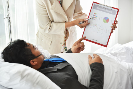 Overworked, Overtime working concepts. Asian Businessman with oxygen mask in grey suit is working while lying on hospital bed with female secretary.の写真素材
