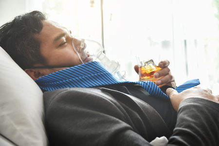 Alcohol addicted businessman with oxygen mask is holding whiskey glass while lying on hospital bed. Sad depressed patient male adult having troubles with alcohol. Asian Drunk man with Alcoholism. Business Failure, Overwoked, Overtime working concepts.の写真素材