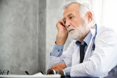 Senior male Doctor is thinking and checking his patient medical records on a clipboard at a Medical room. Stressed. Concentration.の写真素材