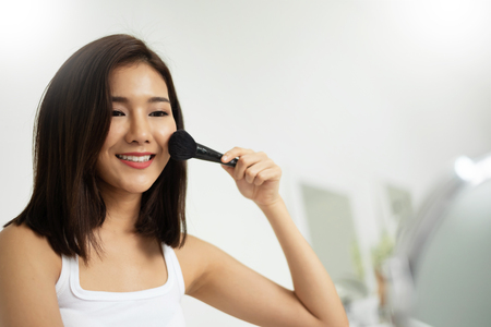 Beautiful Asian woman looking at the mirror and applying makeup with a brush on the cheekbones.の写真素材