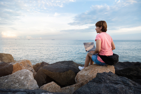 Young Asian Woman Freelance in pink shirt sitting on the rock and working with laptop at outdoor. Sea and sunset background.の写真素材