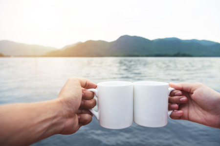 Relaxed Couple hands holding cups of hot coffee with river and mountain view. Enjoying morning. Relax time.の写真素材