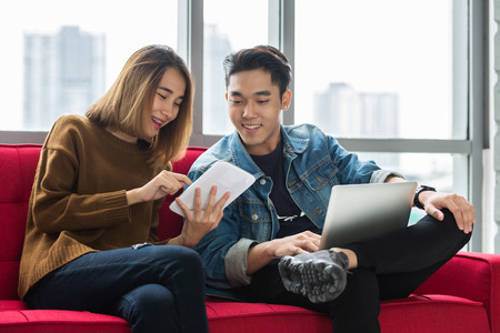 Happy Young couple designer using digital tablet and laptop together. Relaxing University students sitting on red sofa.の写真素材