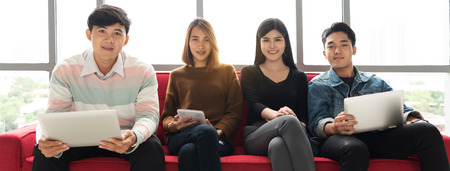 Portrait of Confident designers team holding laptops and digital tablet while sitting on red sofa. Young Business people are looking camera.の写真素材