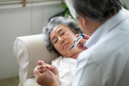Sick Asian Senior Woman laying on couch while her husband holding and looking to temperature....