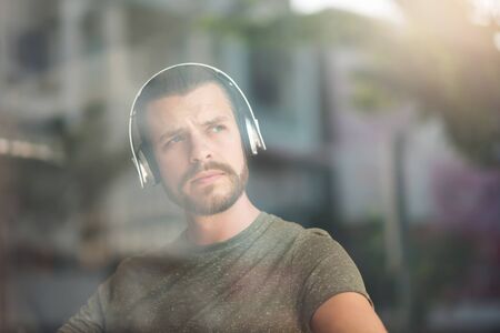 Portrait of bearded man listening to music with headphones. Hipster male looking out at the window...