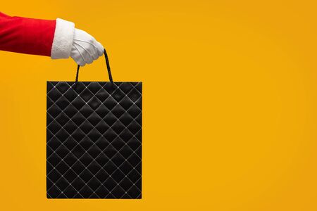Santa Claus hand holding black shopping leather bag on isolated yellow background. Black Friday, Sales, Giving Gift for Christmas and New Year 2019.の写真素材