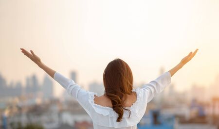 Asian woman raised hands up in the air over city background while standing at outdoor in the morning. Relaxing with Open arms. Rear view.の写真素材