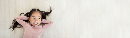 Smiling Asian child girl in pink sweater looking up and sleep on wooden floor on holidays.の写真素材