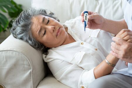 Elderly woman having cold during lie down on couch while Senior man checking temperature of his wife with digital thermometer in the house. Old female having fever. Illness, Disease, Coronavirus, virus, Covid, Spread, Quarantine, Carer.の写真素材