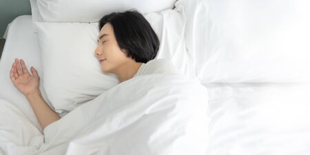 Top view Young man sleeping and dreaming in white bed in the morning. Happy smiling Asian male enjoying lying on comfortable soft bedding in bedroom at home. eyes closed. copy space.の写真素材