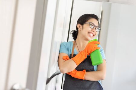 Portrait Happy Young woman wear glasses holding green cleaner spray bottle like at microphone singing in white room. Smiling Asian female in blue shirt with Apron and orange rubber gloves preparing cleaning her house. Having fun, Chores, Houseworkの写真素材