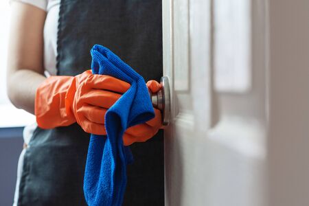 Woman hands in orange rubber gloves cleaning on touching surface of door, doorknob with blue...