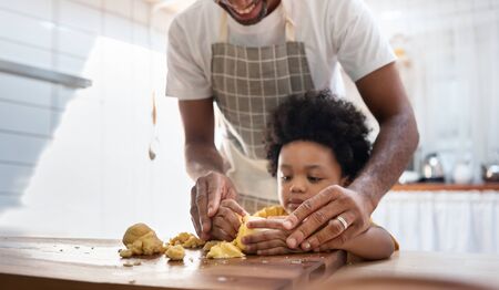 Black family having fun prepare bakery together at home. African American father and adorable son kneading dough in kitchen. Happy loving Man and little helper boy enjoy while making cakes and cookiesの写真素材