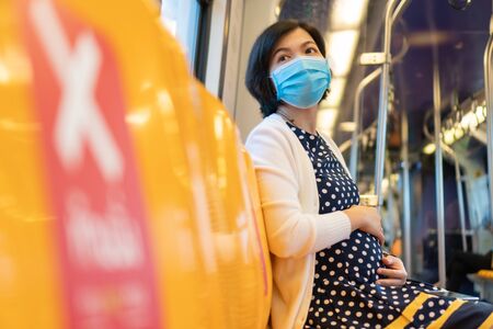 Asian Pregnant Woman wear face mask touch her belly and sit on yellow seat in sky train while go to office. Businesswoman with social distance while pandemic covid-19 or coronavirus. New normal lifeの写真素材