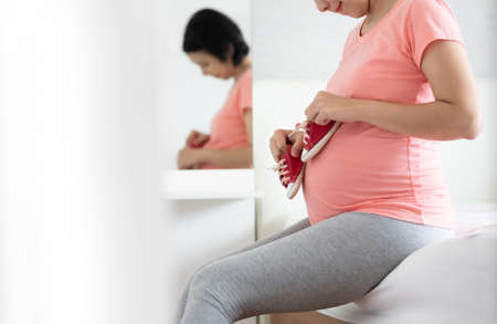 Healthy Pregnant Woman in pink shirt smiling holding Little Baby Red Shoes on Belly. Happy Asian...