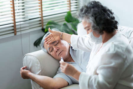 Sick Asian Senior man have flu lying on couch while his Wife in face mask holding and checking...