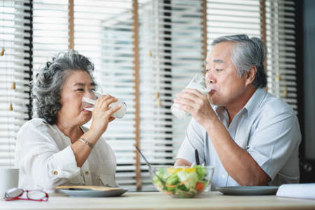 Smiling Asian senior man and woman sitting at table drinking glasses of milk and looking at each...
