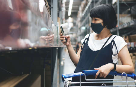 Young Asian Pregnant Woman searching products with mobile phone at mall. Female buyer in protective face mask prevent virus using smartphone while shopping in store during her pregnancy. Self serviceの写真素材
