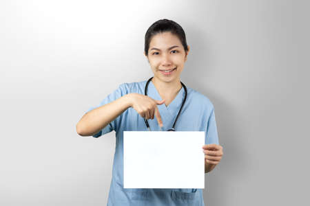 Doctor woman holding white mockup poster isolated on grey background. Physician pointing on blank paper sheet with finger. Portrait friendly nurse wear blue uniform with happy face and confident smileの写真素材