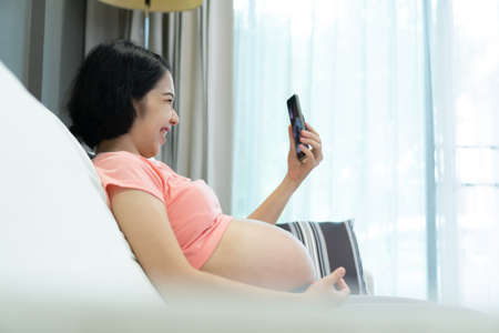 Attractive Asian Pregnant woman in pink shirt making video call online for talking with family on...