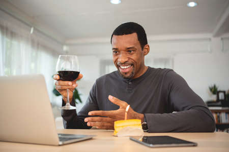 Happy smiling Black male doing online celebration with toasting red wine via video call on laptop during quarantine lockdown. African American man is meeting with his friends birthday togetherの写真素材