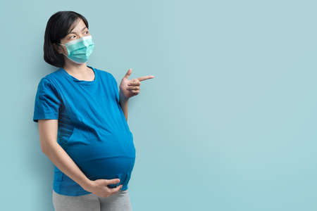 Happy Smiling Asian Pregnant woman wearing protective face mask and blue t-shirt pointing and looking to copy space. Female standing touching on belly over isolated background.の写真素材