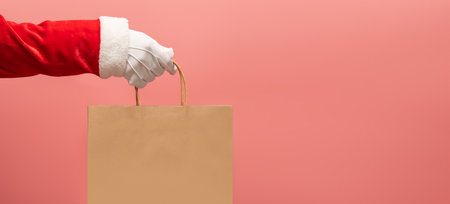 Santa Claus hand holding brown shopping paper bag on pink isolated background. Banner and copy spaceの写真素材