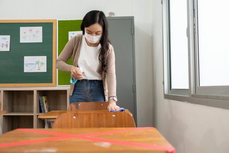 Asian Woman Teacher wearing protective face mask cleaning disinfecting with antibacterial alcohol spray bottle on table surface in classroom before reopening school.の写真素材