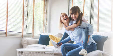 Happy Beautiful Mother carrying or piggyback her little daughter laughing playing and having fun together on sofa at home.の写真素材