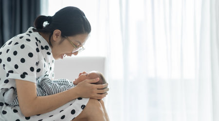 Happy Asian Young mom sitting on bed playing with small infant and laughing together. Loving Female enjoying embracing her son in her hands. Mother and little baby Bonding and Familyの写真素材