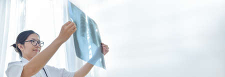 Smiling Asian Young Female doctor holding looking and examining X-ray film of her patient in medical...