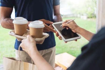 Close up of Asian Woman using pen signing digital signature on mobile tablet after receive hot drink or coffee from African American delivery man in blue uniform at door of home.の写真素材