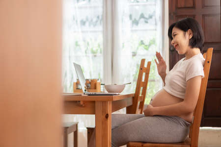 Asian Pregnant woman making online video call on laptop at home. Smiling Young Mother talking waving hand to her family on notebook while her pregnancy.の写真素材